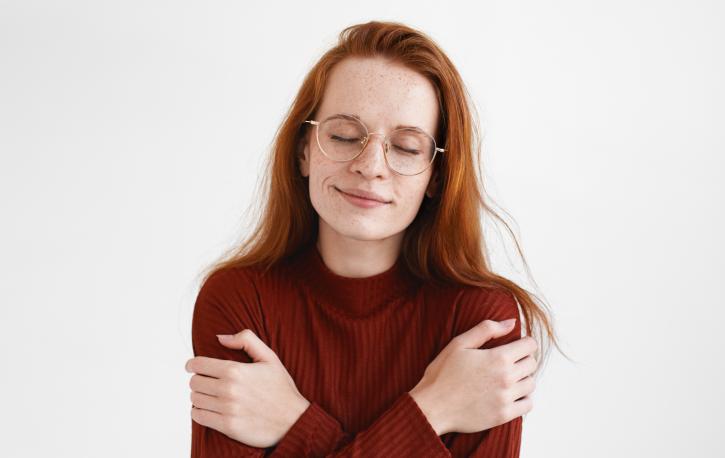 Eine Frau mit Brille und rötlichen Haaren umarmt sich selbst.