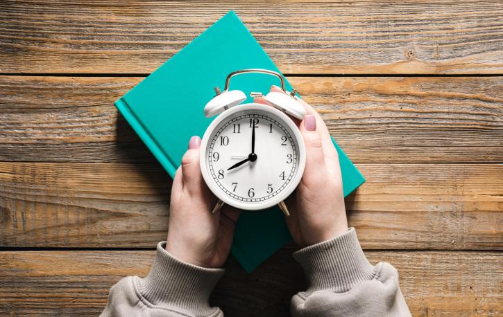 Man sieht einen weißen Wecker von oben, der von zwei Händen umfasst wird.