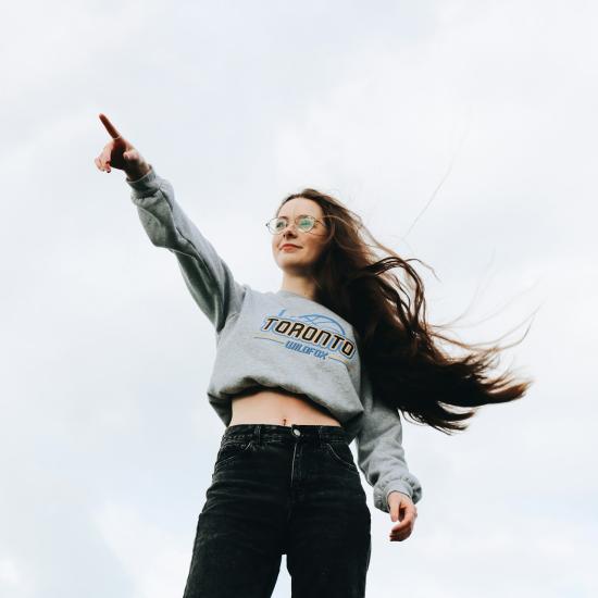 Eine junge Frau zeigt selbstbewusst in die Ferne, im Hintergrund ein bewölkter Himmel, ihre Haare flattern im Wind.