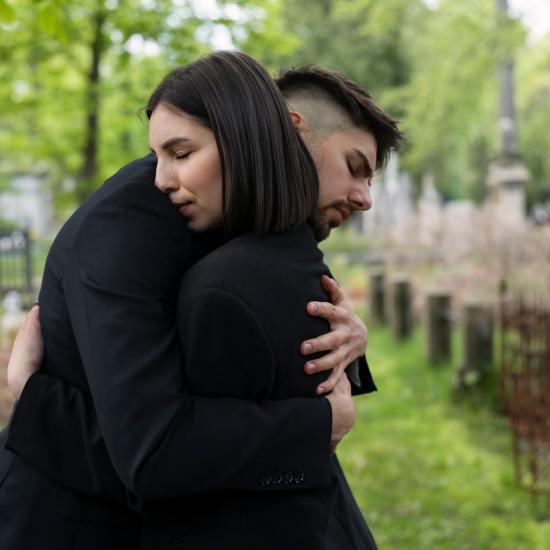 Ein Mann und eine Frau in schwarzer Kleidung umarmen sich auf einem Friedhof.