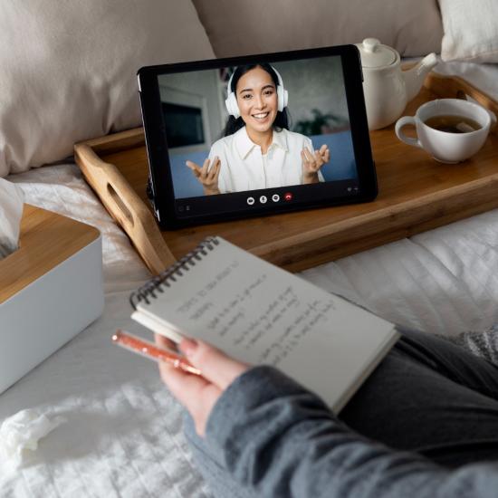 Man sieht die Hände einer Person, die einen Notizblock in der Hand hält und auf dem Bett vor einem Tablet sitzt.