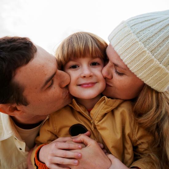 Ein Kind zwischen seinen Eltern, die ihm je einen Kuss auf die Wange geben.