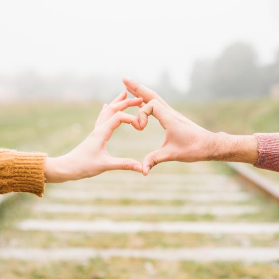 Die Hände von zwei Personen formen ein Herz, auf dem Boden sieht man Bahngleise.