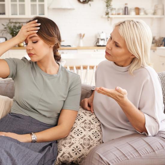 Eine junge und eine mittelalte Frau sitzen nebeneinander auf einem Sofa und unterhalten sich angestrengt.