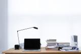 Frontansicht eines Schreibtischs mit Lampe, Laptop, Büchern und Dokumenten. Im Hintergrund eine weiße Wand oder ein Fenster mit sehr hellen Gardinen.