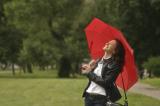 Eine weiße Frau steht lachend auf einer Wiese. Sie hält sich einen geöffneten roten Schirm über den Kopf. Im Hintergrund ein Baum.