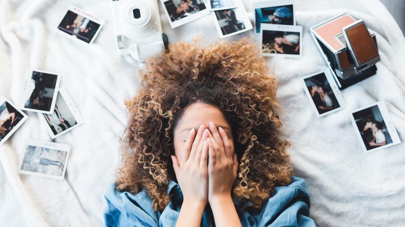Junge Frau liegt auf dem Rücken auf dem Bett und hält sich die Hände vor das Gesicht. Um ihren Kopf herum sind Polaroidfotos ausgebreitet.
