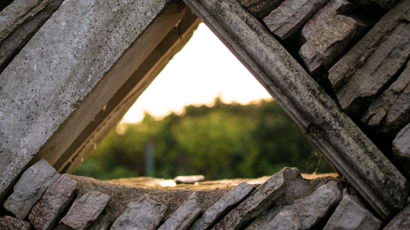 Steinmauer, durch die Lücke in Form eines gleichschenkligen Dreiecks sieht man Sonnenlicht und grüne Bäume.