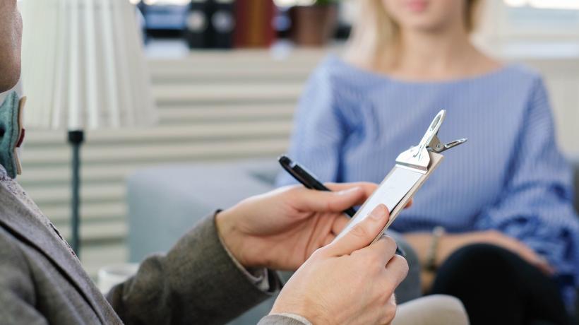 Ein Psychologe sitzt bei einer Patientin und hält ein Klemmbrett in der Hand.