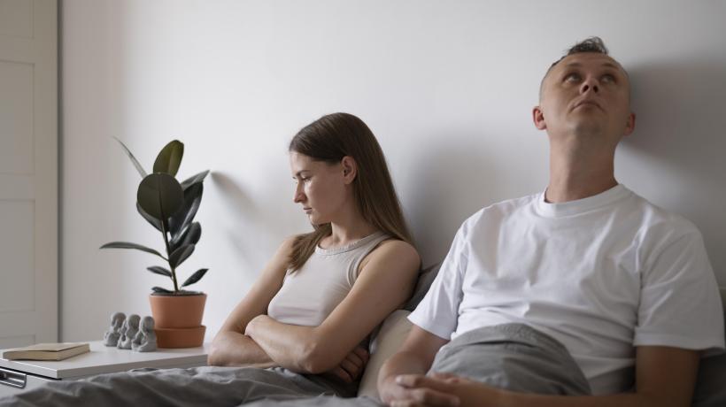 Ein junges Pärchen sitzt auf dem Bett und schaut missgestimmt in unterschiedliche Richtungen.