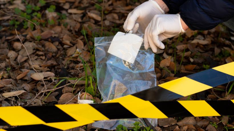 Man sieht zwei behandschuhte Hände, die hinter einem Absperrband etwas in eine Plastiktüte tun.