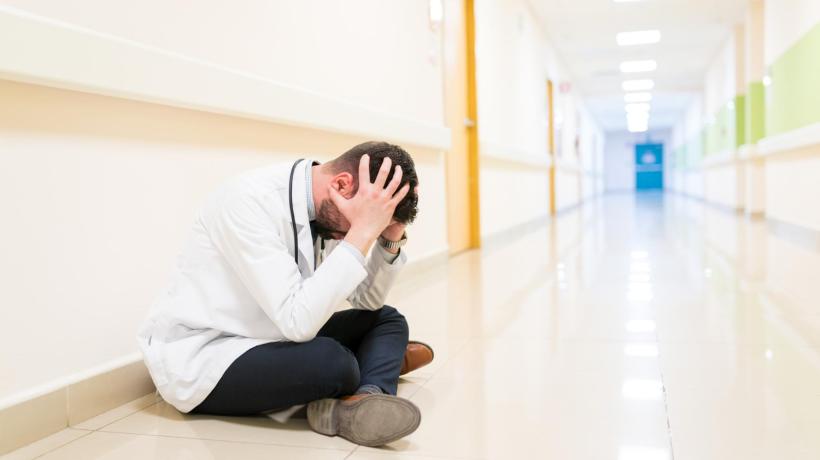 Ein Mann in medizinischem Kittel sitzt mit in die Hände gestütztem Kopf auf einem Krankenhausflur.