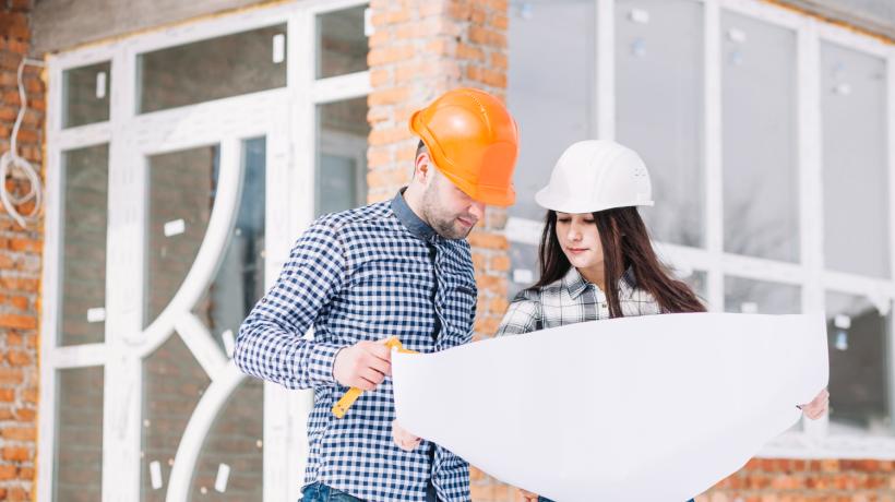 Ein Mann und eine Frau mit Bauhelm stehen vor einem Haus und gucken in einen großen Plan.