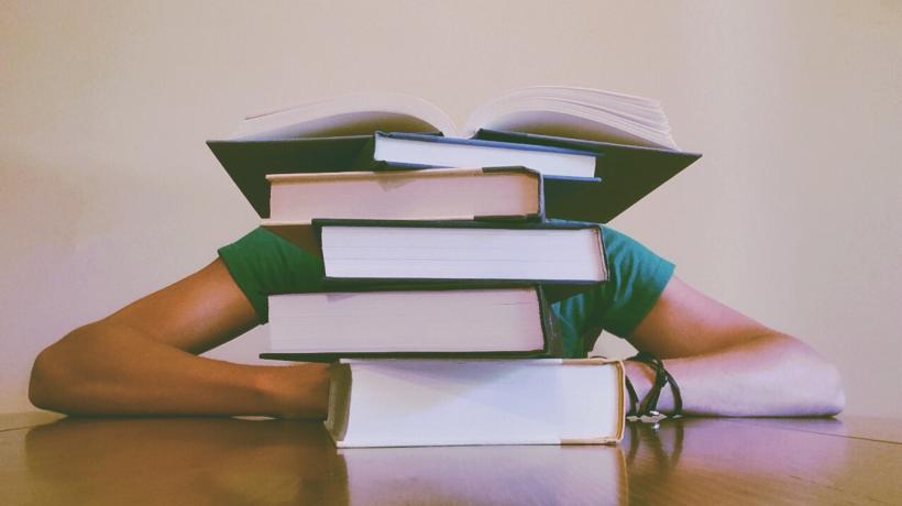 Person verzweifelt hinter Bücherstapel mit Kopf auf dem Tisch.