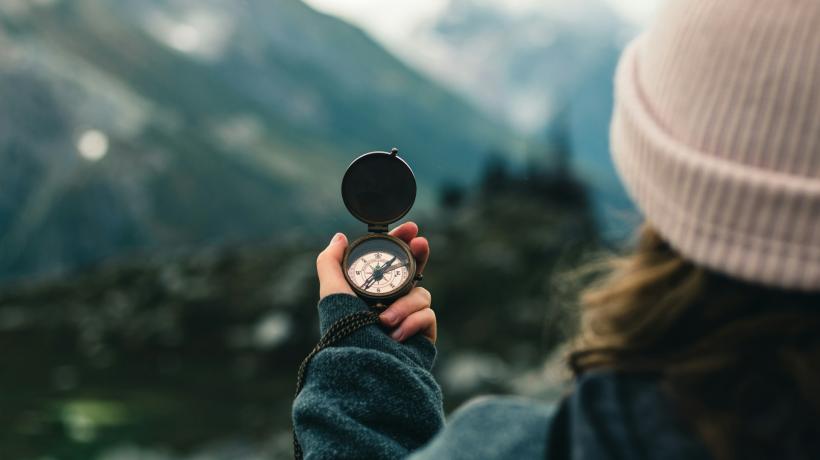 Man sieht eine Frau von hinten, die einen Kompass in die Höhe hält; im Hintergrund eine Berglandschaft