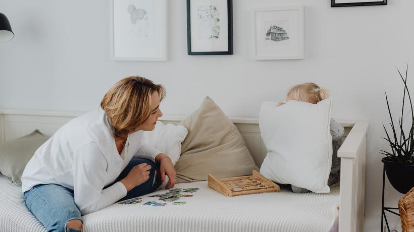 Eine Mutter sitzt auf der Couch und versucht spielerisch, ihre Tochter hervorzulocken, die sich hinter einem Kissen versteckt hat.
