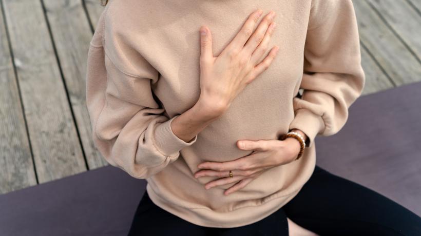 Man sieht den Oberkörper einer Frau in cremefarbenem Pullover, die eine Hand auf die Brust, die andere auf den Bauch gelegt hat.