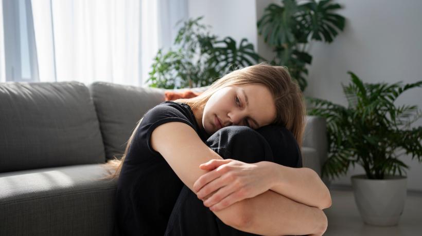 Eine junge Frau hat die Arme um ihre Beine geschlungen und sitzt betrübt vor ihrem Sofa.