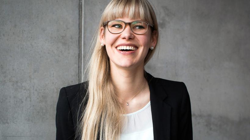 Porträtaufnahme von Eva Asselmann, sie hat lange blonde Haare und einen Pony, trägt eine Brille, ein weißes Shirt und eine schwarze Jacke.