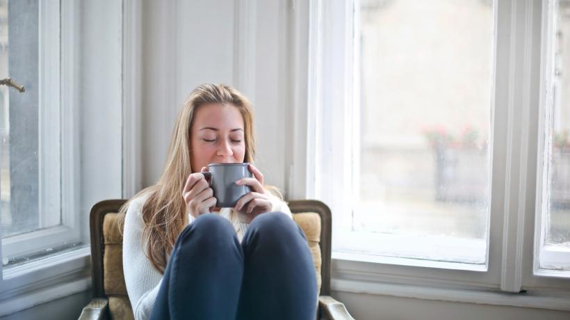 Eine Frau sitzt mit angezogenen Beinen und zufriedenem Gesichtsausdruck mit einer Tasse auf einem Stuhl.