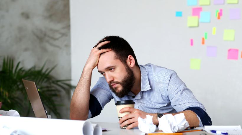 Ein Mann sitzt mit Kaffeebecher in der Hand und aufgestütztem Kopf missmutig am Schreibtisch