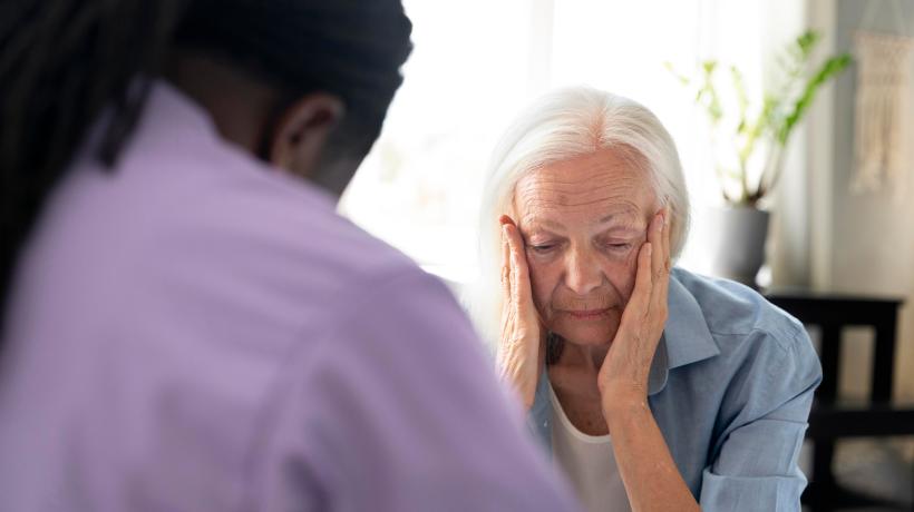 Eine ältere Patientin und ihre Psychotherapeutin sitzen sich gegenüber, die Patientin schaut belastet zu Boden.