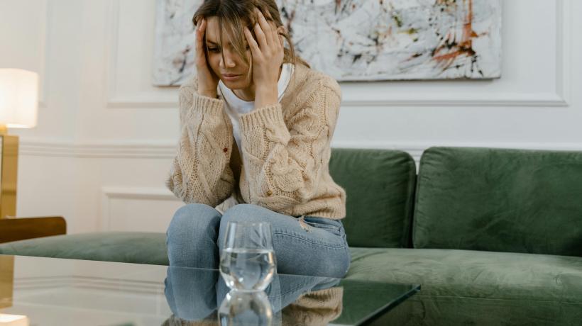 Eine junge Frau sitzt auf einem Sofa, hält sich die Hände an die Schläfen und schaut betrübt zu Boden.