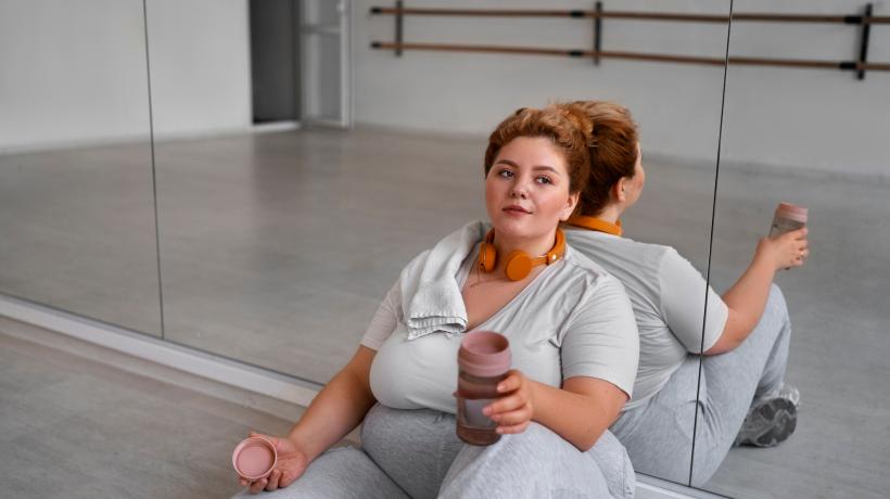 Eine mehrgewichtige Frau lehnt an einer Spiegelwand im Tanzstudio und hält eine Trinkflasche in der Hand.