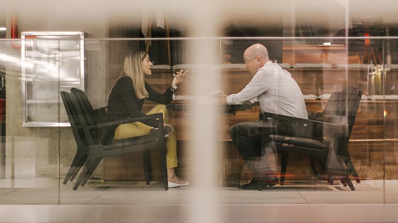 Blick durch ein Fenster in Raum. Ein Mann und eine Frau sitzen sich gegenüber am Tisch, reden.