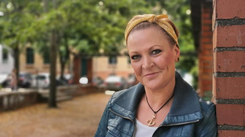 Martina Leisten lehnt an einer Hauswand und lächelt verschmitzt in die Kamera. Sie trägt ein gelbes Haarband, eine blaue Jeansjacke und ein weißes Shirt, dazu eine Kette. Im Hintergrund sind ein Fußweg, Bäume und Autos zu sehen.