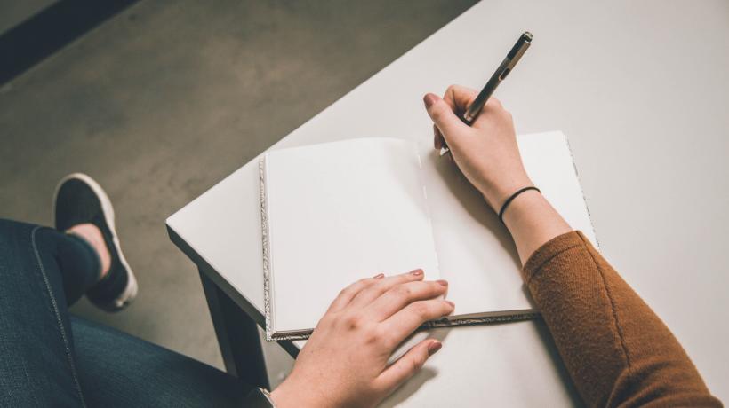 Person sitzt am Schreibtisch mit Stift in der Hand und schreibt in leeres Notizbuch.