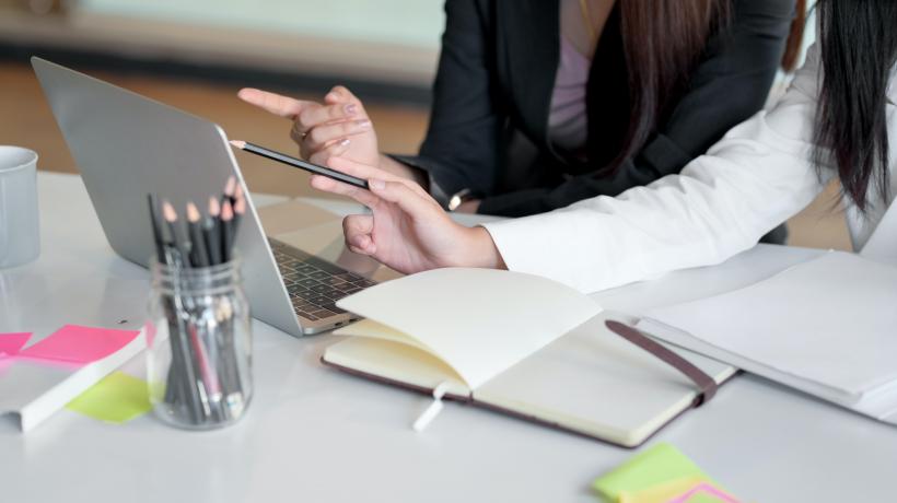 Zwei Frauen sitzen am Schreibtisch und zeigen mit dem Zeigefingerauf einen Laptop.