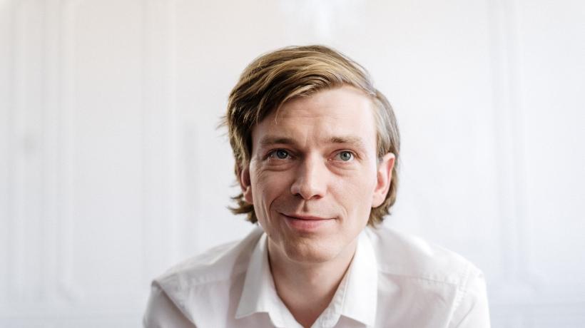 Portrait eines blonden Mannes der schmunzelt, al sei ihm gerade ein guter Einfall gekommen.