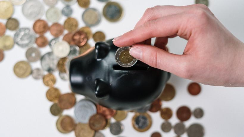 Man sieht eine Hand, die Geld in ein schwarzes Sparschwein wirft.