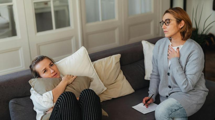 Eine Frau liegt ein Kissen umarmend auf dem Sofa, neben ihr sitzt eine Frau mit Notizzettel
