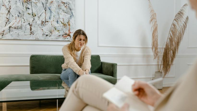 Eine Frau hockt in sich zusammengesackt auf einem Sofa in einem Therapiezimmer, ihr gegenüber sitzt eine Person, die Unterlagen ausfüllt.