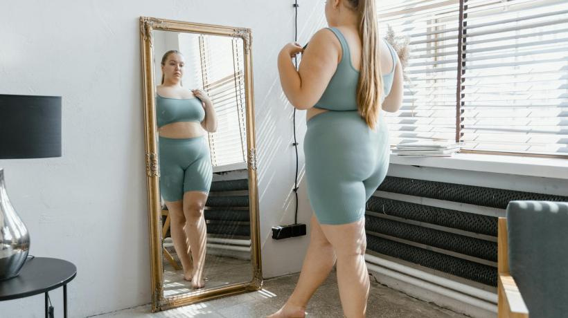 Eine übergewichtige junge Frau in kurzer Hose und Top steht vor einem Spiegel.
