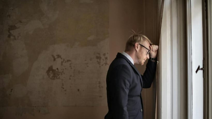 Ein Mann in Anzug lehnt mit aufgestützter Stirn neben einem Fenster an der Wand.