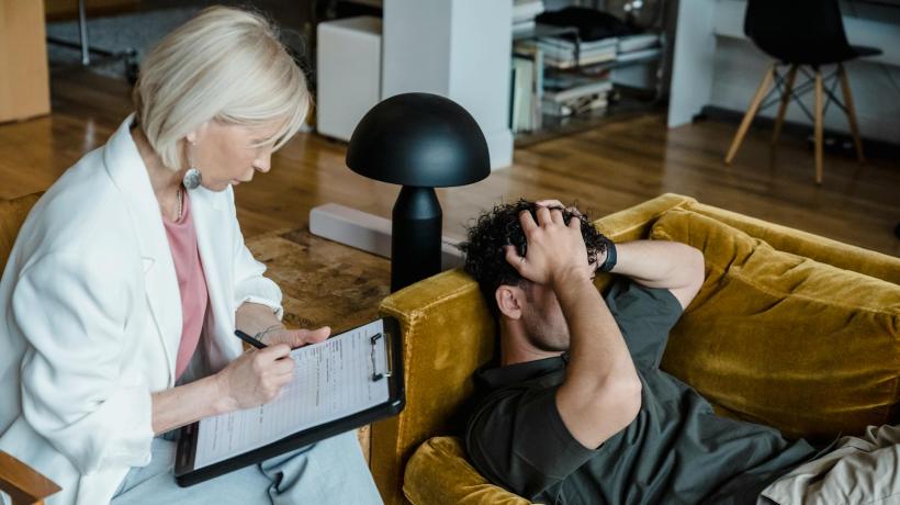 Ein Mann liegt auf der Couch seiner Therapeutin und hält sich angestrengt den Kopf. Die Therapeutin schreibt auf ein Klemmbrett.