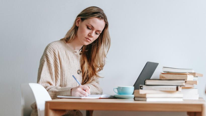 Eine junge Frau sitzt konzentriert am Tisch und schreibt auf einem Block, vor ihr stehen Laptop, eine Tasse und ein Stapel Bücher.