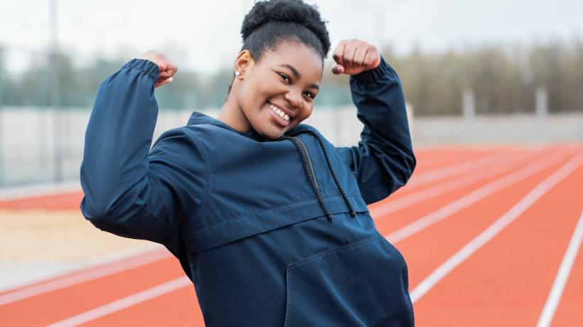 Eine junge Frau steht lächelnd an einer Outdoor-Sportanlage und reckt triumphierend die Hände in die Höhe