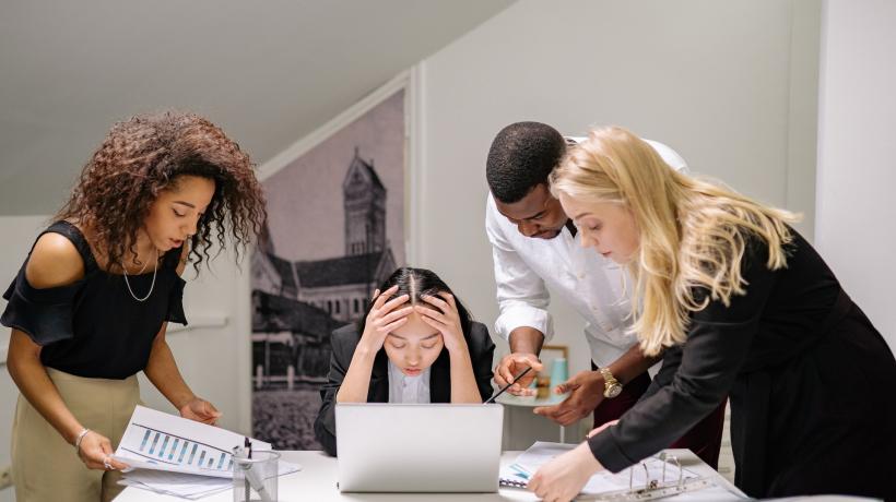 Eine jugen Frau sitzt vorm Laptop und schlägt die Hände über dem Kopf zusammen, während zwei Kolleginnen und ein Kollege ihr immer mehr Unterlagen und Arbeit auf den Tisch legen.