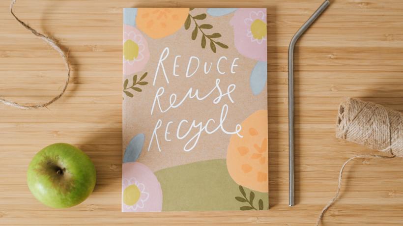 Von oben blickt man auf eine Tischplatte, auf der akurat angeordnet ein Notizbuch mit der Aufschrift "Reduce, Reuse, Recycle" sowie ein Apfel, ein nachhaltiger Strohhalm aus Metall und eine Rolle Bindfaden liegen.