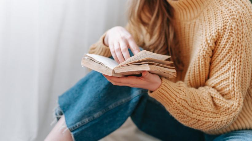 Eine Frau in Jeans und Pull sitzt auf dem Boden und liest ein Buch.
