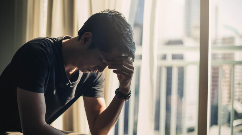 Ein Mann sitzt nach vorn gebeugt am Fenster und stützt seine Stirn auf seine linke Hand.