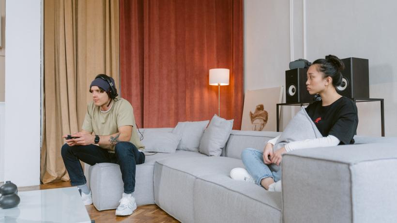 Ein junger Mann sitzt mit Kopfhörern und Controller in den Händen auf der Couch und blickt gebannt nach vorn, am anderen Ende der Couch sitzt eine junge Frau, die ihn besorgt ansieht.
