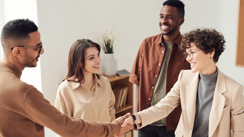 Zwei junge Männer und zwei junge Frauen im legeren Businesslook stehen beieinander, eine der Frauen und einer der Männer geben sich lächelnd die Hand.