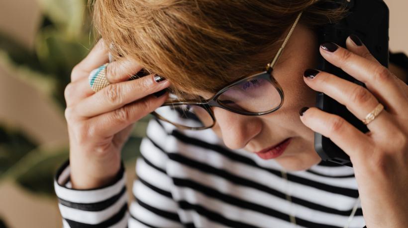 Eine Frau mit Brille, kurzen Haaren und ernstem Gesichtsausdruck ist am Telefon und greift sich mit der freien Hand an den Kopf.