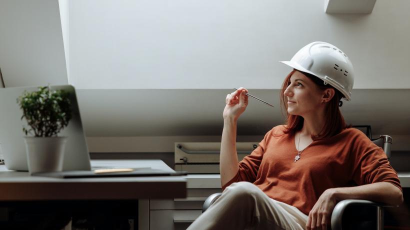 Eine Frau sitzt mit Bauhelm und Stift an einem Tisch und blickt sinnierend in die Luft.