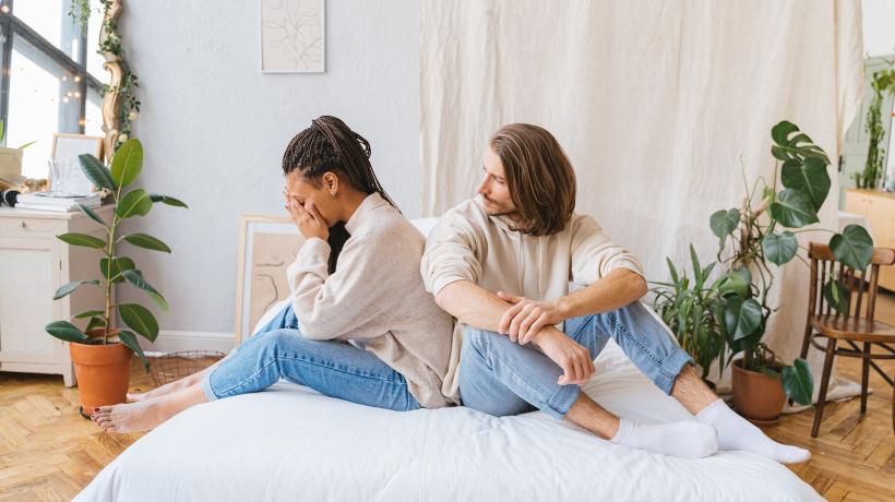 Eine Frau und ein Mann sitzen angezogen auf dem Bett, sie hat ihm den Rücken zugedreht und hält die Hände vor ihrem Gesicht, er schaut fragend zu ihr rüber.
